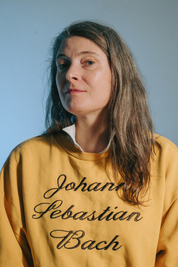 eine Person mit langem Haar und einem gelben Pullover auf "Johann Sebastian Bach" steht, schaut in die Kamera