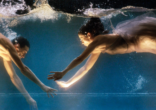 Zwei Menschen schwimmen sich unter Wasser entgegen. Sie sind angeleuchtet von unten.