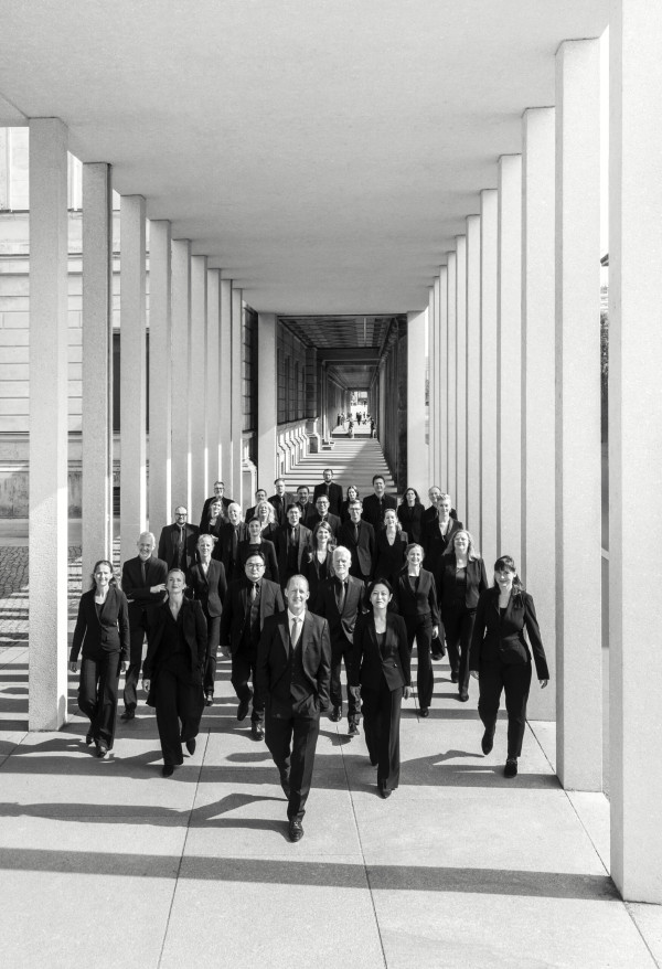 Eine gruppe Menschen in schwarzer Kleidung läuft in einem Säulengang in Richtung der Kamera. Das Licht bricht durch die Säulen